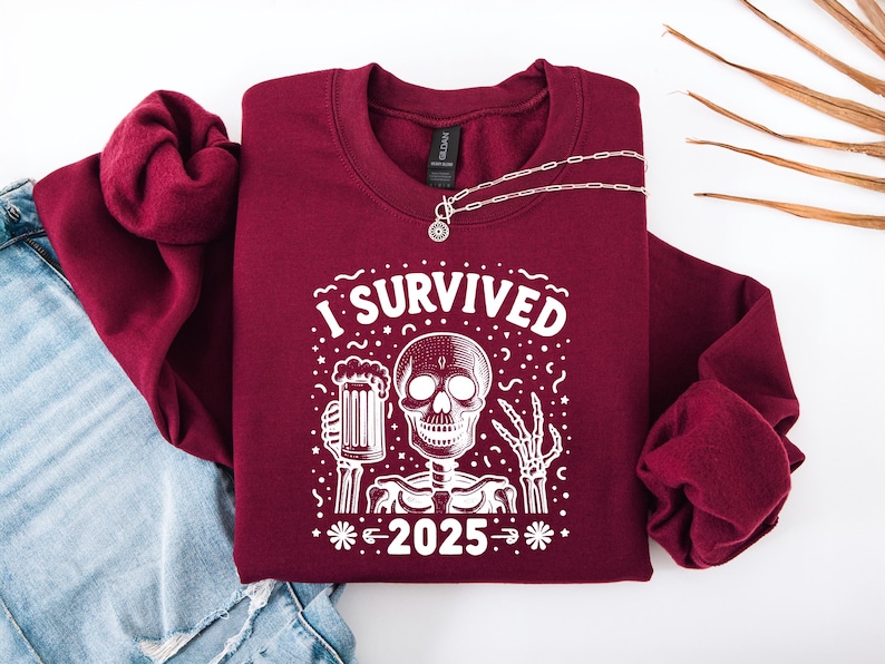 Puede incluir: Sudadera burdeos con el texto blanco "I SURVIVED 2025". El gr&aacute;fico presenta un esqueleto sosteniendo una jarra de cerveza. Un collar plateado con un colgante est&aacute; sobre la sudadera. La sudadera est&aacute; sobre unos vaqueros.