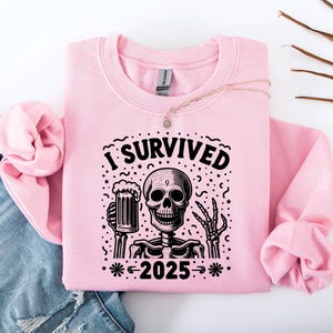 Puede incluir: Sudadera rosa claro con un gr&aacute;fico negro de un esqueleto sosteniendo una jarra de cerveza. El texto "I SURVIVED 2025" est&aacute; encima y debajo del esqueleto. La sudadera est&aacute; doblada sobre unos vaqueros azules.