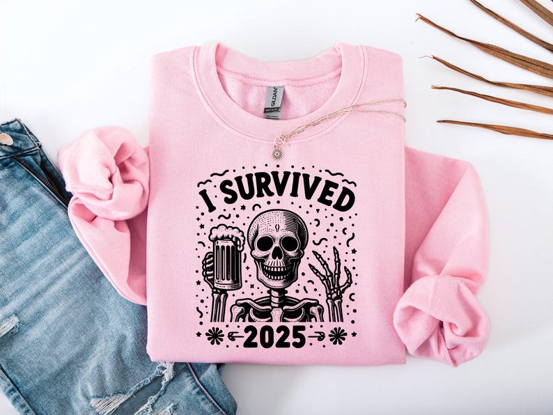 Puede incluir: Sudadera rosa claro con un gr&aacute;fico negro de un esqueleto sosteniendo una jarra de cerveza. El texto "I SURVIVED 2025" est&aacute; encima y debajo del esqueleto. La sudadera est&aacute; doblada sobre unos vaqueros azules.