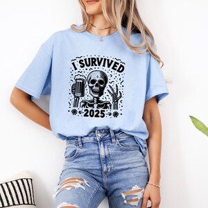 Puede incluir: Camiseta azul claro con un dise&ntilde;o negro de un esqueleto sosteniendo una jarra de cerveza. El texto "I Survived 2025" est&aacute; impreso encima y debajo del esqueleto. La persona lleva vaqueros azules.