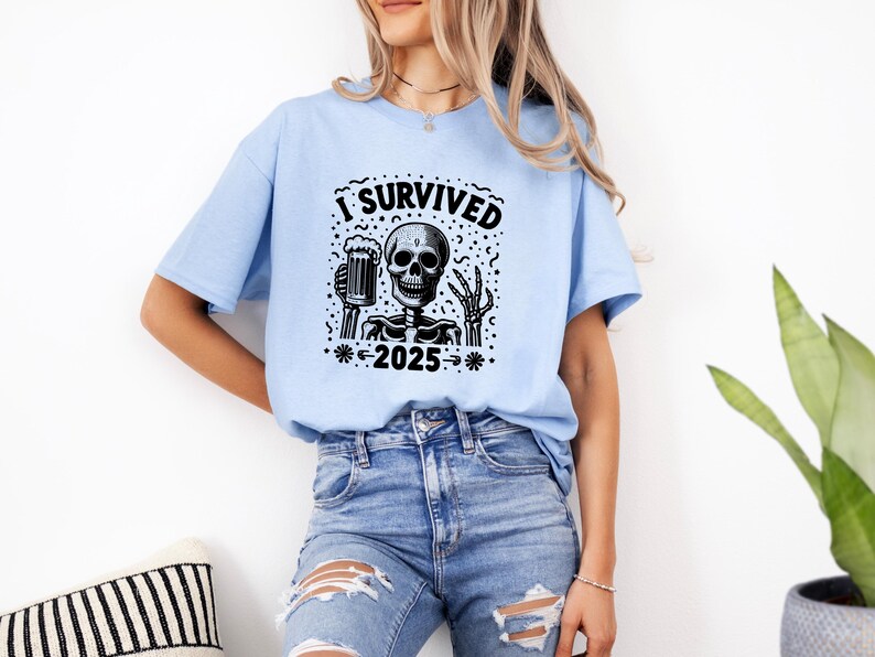 Puede incluir: Camiseta azul claro con un dise&ntilde;o negro de un esqueleto sosteniendo una jarra de cerveza. El texto "I Survived 2025" est&aacute; impreso encima y debajo del esqueleto. La persona lleva vaqueros azules.
