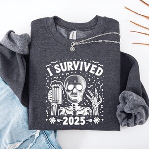 Puede incluir: Sudadera gris oscuro con un gr&aacute;fico blanco de un esqueleto sosteniendo una jarra de cerveza y el texto "I SURVIVED 2025". Un collar plateado est&aacute; sobre la sudadera. La sudadera est&aacute; doblada sobre unos vaqueros azules.