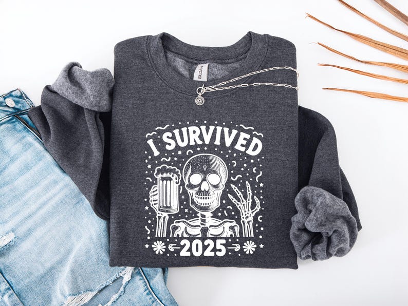 Puede incluir: Sudadera gris oscuro con un gr&aacute;fico blanco de un esqueleto sosteniendo una jarra de cerveza y el texto "I SURVIVED 2025". Un collar plateado est&aacute; sobre la sudadera. La sudadera est&aacute; doblada sobre unos vaqueros azules.