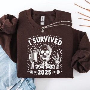 Puede incluir: Una sudadera marr&oacute;n oscuro con el texto "I SURVIVED 2025" y un dise&ntilde;o de esqueleto. El esqueleto sostiene una jarra de cerveza. Un collar plateado con un colgante redondo est&aacute; sobre la sudadera. Se ven unos vaqueros azules.
