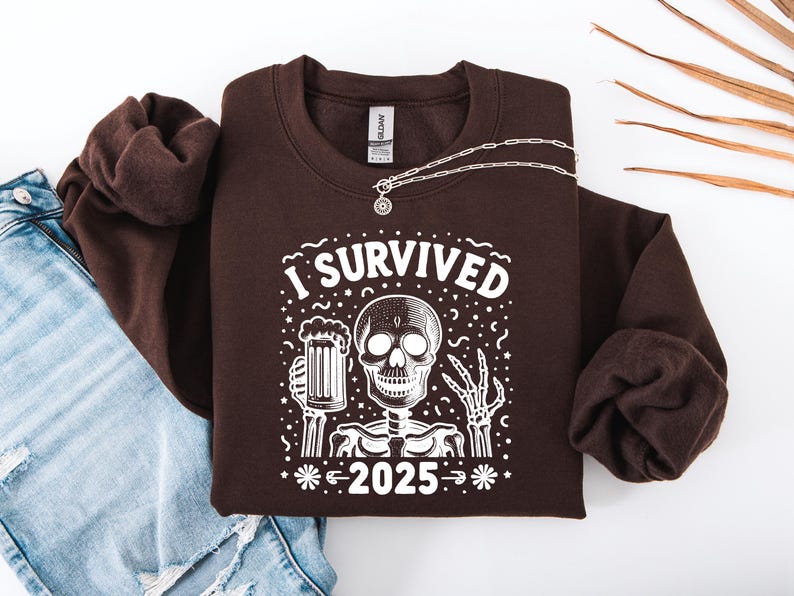 Puede incluir: Una sudadera marr&oacute;n oscuro con el texto "I SURVIVED 2025" y un dise&ntilde;o de esqueleto. El esqueleto sostiene una jarra de cerveza. Un collar plateado con un colgante redondo est&aacute; sobre la sudadera. Se ven unos vaqueros azules.