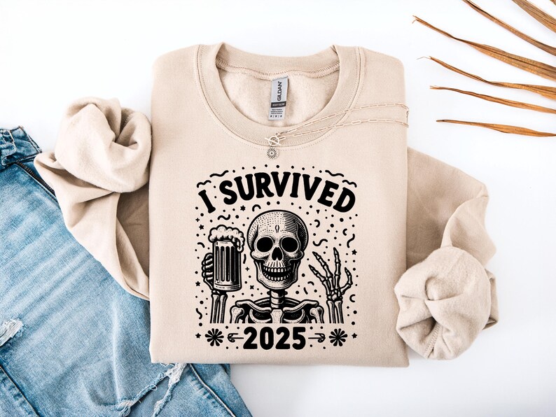 Puede incluir: Sudadera beige con un gr&aacute;fico negro de un esqueleto sosteniendo una jarra de cerveza, con el texto "I Survived 2025". La sudadera est&aacute; doblada sobre unos vaqueros azules.