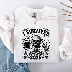 Puede incluir: Sudadera blanca con un gr&aacute;fico en blanco y negro de un esqueleto sosteniendo una jarra de cerveza. El texto "I Survived 2025" est&aacute; encima del esqueleto. La sudadera est&aacute; doblada sobre unos vaqueros azules.