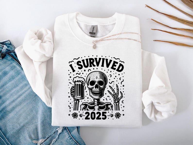 Puede incluir: Sudadera blanca con un gr&aacute;fico en blanco y negro de un esqueleto sosteniendo una jarra de cerveza. El texto "I Survived 2025" est&aacute; encima del esqueleto. La sudadera est&aacute; doblada sobre unos vaqueros azules.