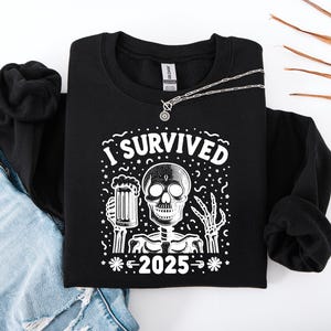 Puede incluir: Sudadera negra con un gr&aacute;fico blanco que dice "I Survived 2025" con un esqueleto sosteniendo una cerveza. Un collar plateado con un colgante redondo est&aacute; sobre la sudadera. Se ven unos vaqueros azules en el fondo.