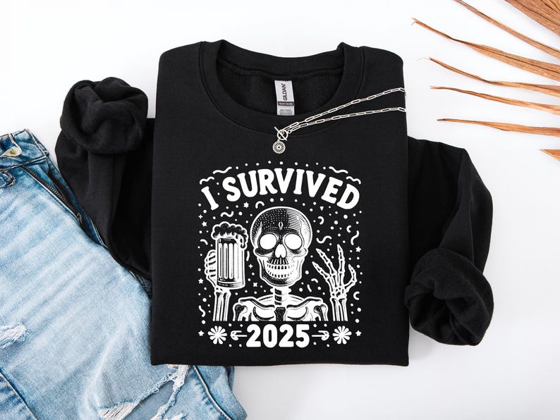 Puede incluir: Sudadera negra con un gr&aacute;fico blanco que dice "I Survived 2025" con un esqueleto sosteniendo una cerveza. Un collar plateado con un colgante redondo est&aacute; sobre la sudadera. Se ven unos vaqueros azules en el fondo.