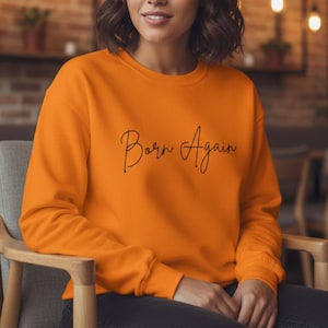 May include: Bright orange sweatshirt featuring the words "Born Again" in black cursive script. The person is wearing the sweatshirt. The sweatshirt is a solid orange colour.