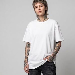 Pode incluir: Uma camiseta branca de manga curta. A camisa é de cor lisa e parece ser feita de um material macio. A modelo está usando jeans pretos. A modelo tem tatuagens nos braços e pescoço.
