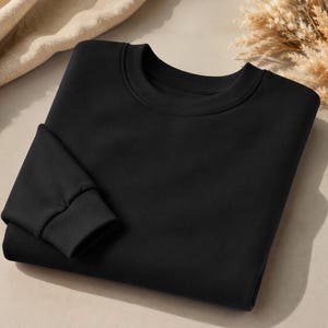 Puede incluir: Una sudadera negra lisa de manga larga con cuello redondo cl&aacute;sico y pu&ntilde;os acanalados. La prenda est&aacute; doblada cuidadosamente, mostrando una textura de tela suave y uniforme en toda la parte delantera y en la secci&oacute;n visible de la manga.