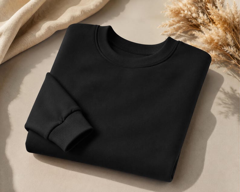 Puede incluir: Una sudadera negra lisa de manga larga con cuello redondo cl&aacute;sico y pu&ntilde;os acanalados. La prenda est&aacute; doblada cuidadosamente, mostrando una textura de tela suave y uniforme en toda la parte delantera y en la secci&oacute;n visible de la manga.