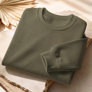 Puede incluir: Una sudadera de cuello redondo en color verde oliva s&oacute;lido, con mangas largas de pu&ntilde;os acanalados y un cuello tambi&eacute;n acanalado. La prenda est&aacute; confeccionada en un tejido liso y opaco, con una textura interior similar al forro polar.