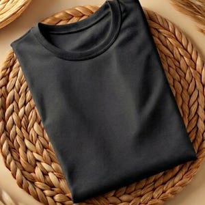 May include: A folded, solid black t-shirt with a crew neck. The shirt is neatly arranged on a woven, circular placemat, creating a simple, yet visually appealing presentation. The fabric appears soft and smooth.