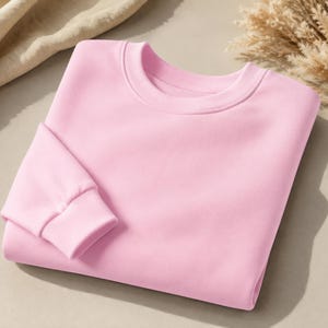 Puede incluir: Una sudadera de cuello redondo en color rosa claro s&oacute;lido, con mangas largas que presentan pu&ntilde;os acanalados y un cuello tambi&eacute;n acanalado. La prenda est&aacute; doblada cuidadosamente, mostrando una textura de tela lisa y uniforme en toda su superficie.
