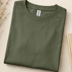 Puede incluir: Una camiseta verde oliva, doblada cuidadosamente, con cuello redondo y mangas cortas. La etiqueta de la marca Gildan es visible en el cuello. Un libro cerrado y espigas de trigo secas en el fondo, creando una estética minimalista.