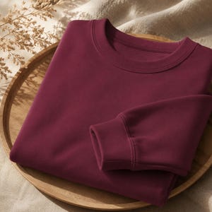 Puede incluir: Una sudadera de cuello redondo en color burdeos profundo s&oacute;lido, con mangas largas de pu&ntilde;os acanalados y un cuello a juego tambi&eacute;n acanalado. La prenda est&aacute; doblada cuidadosamente, mostrando una textura de tela suave y uniforme.