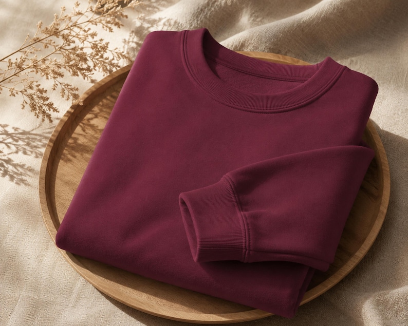 Puede incluir: Una sudadera de cuello redondo en color burdeos profundo s&oacute;lido, con mangas largas de pu&ntilde;os acanalados y un cuello a juego tambi&eacute;n acanalado. La prenda est&aacute; doblada cuidadosamente, mostrando una textura de tela suave y uniforme.