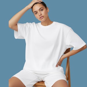 Puede incluir: Un conjunto de camiseta y pantalón corto blancos. La camiseta tiene mangas cortas y cuello redondo. El pantalón corto es de color blanco a juego. La persona está sentada en una silla de madera sobre un fondo azul.