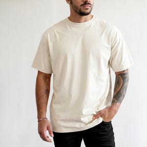 Könnte beinhalten: Ein cremefarbenes Kurzarm-T-Shirt mit Rundhalsausschnitt. Das Kleidungsstück hat eine lockere Passform, einen geraden Saum und ein schlichtes, musterloses Design. Der Stoff wirkt glatt und blickdicht und sorgt für einen sauberen, einfachen Look.