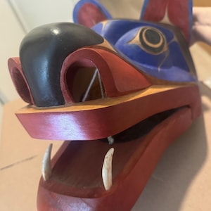 Pode incluir: Uma máscara de madeira esculpida que retrata uma cabeça de animal, com um focinho preto, orelhas azuis e vermelhas e uma boca vermelha com dois dentes visíveis. A máscara apresenta um design detalhado com várias cores e formas, mostrando o artesanato tradicional.