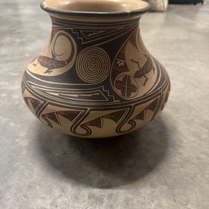 May include: A tan-colored ceramic vase with a wide body and a narrow neck. The vase is decorated with geometric patterns and stylized animal figures in black and red. The rim is dark brown.