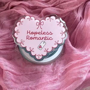 May include: A heart-shaped acrylic charm with a scalloped edge, featuring a light pink background, a small bow graphic, and the text "Hopeless Romantic" in dark pink script, accompanied by a small white tag icon with a pink heart.