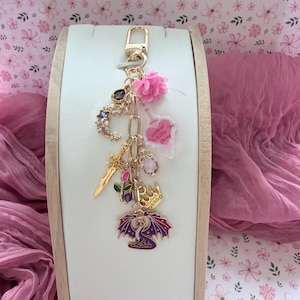 May include: A gold-coloured keychain featuring various charms: a crescent moon, a sword, a crown, and a purple dragon. It also includes a pink flower and a heart-shaped charm with the text "violent little thing". The background displays a floral pattern.