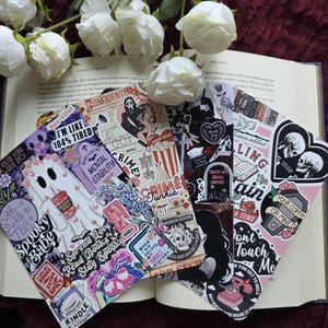 May include: A selection of decorative bookmarks with a gothic and spooky aesthetic, featuring sticker designs with phrases such as "Crime Junkie" and "Spooky Babe." White roses and an open book are visible.