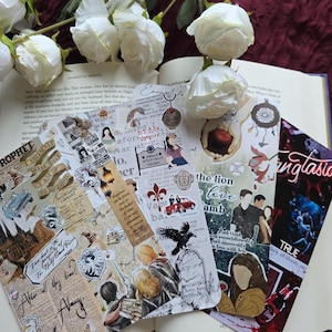 May include: A selection of decorative bookmarks with diverse designs, including text and illustrations. The bookmarks are displayed on an open book, with white roses in the background. The bookmarks feature themes like fantasy and romance.