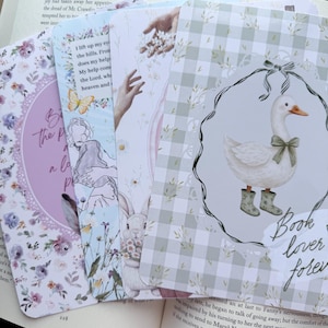 Peut inclure: Ensemble de marque-pages décoratifs avec des illustrations florales, de lapin et d'oie. Les marque-pages présentent des couleurs pastel et le texte "Book lover forever". Ils sont placés sur un livre ouvert.