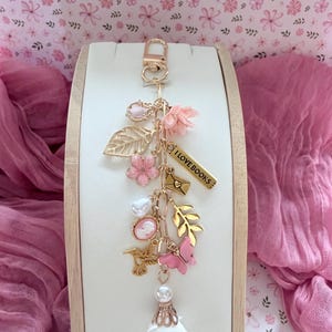 Pode incluir: Um chaveiro dourado com vários berloques: flores cor-de-rosa, uma borboleta, um camafeu e uma folha. O chaveiro inclui um berloque com o texto "I LOVE BOOKS". Uma borla branca e uma pérola completam o design.
