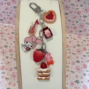 Könnte beinhalten: Ein silberfarbener Schlüsselanhänger mit mehreren Miniatur-Anhängern, darunter ein rosa Cartoon-Hase, ein Erdbeertörtchen, ein Glas Erdbeermarmelade, ein Erdbeergetränk, eine realistische Erdbeere, ein rosa Gummibärchen und ein Erdbeerschichtkuchen.
