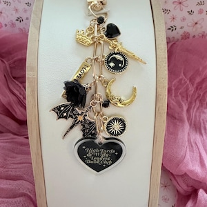 May include: A gold-coloured keychain with various charms, including a crown, heart, moon, and bat. The keychain features a heart-shaped charm with the text "Highlords & Wing Leaders Book Club". The background is pink.