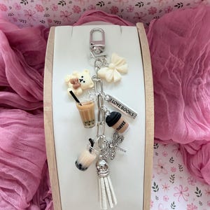 Op de afbeelding: Een zilveren sleutelhanger met meerdere bedels, waaronder een teddybeer, een strik, een koffiekopje en een libelle. De sleutelhanger heeft ook een klein label met de tekst "I LOVE BOOKS". De bedels zijn in verschillende kleuren, waaronder wit, bruin en zwart.