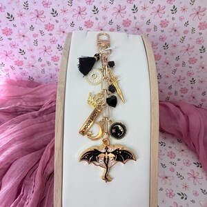 May include: A gold-colored keychain with multiple charms, including a black tassel, a crown, a crescent moon, and a dragon. The keychain also features a charm that reads "I LOVE BOOKS". The background is a floral pattern.