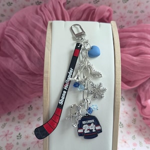 Peut inclure: Un porte-clés argenté avec une breloque bâton de hockey noir et rouge portant le texte Shane Hollander, un cœur bleu, un patin à glace argenté, un flocon de neige, une figurine de joueur de hockey et un maillot bleu marine avec Hollander 24.