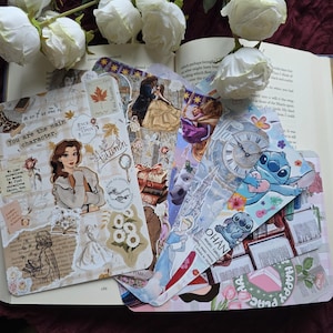 Puede incluir: Varios marcadores decorativos con diversos diseños, incluyendo personajes de dibujos animados y patrones florales, se muestran en un libro abierto. Rosas blancas en el fondo. El texto "You are the main character" es visible. Ideales para amantes de la lectura.