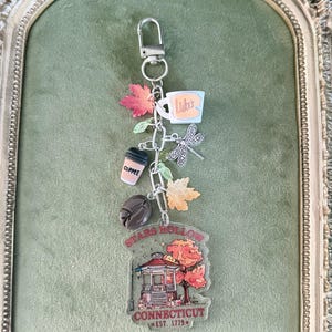 May include: A silver-toned keyring featuring a metal lobster clasp and a chain adorned with charms including a white mug labelled Luke's, a coffee cup, a coffee bean, a dragonfly, and two maple leaves, ending in a Stars Hollow Connecticut charm.