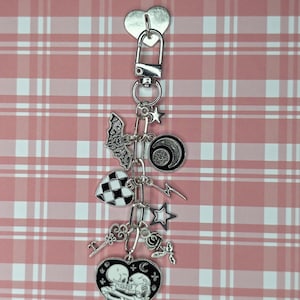 Op de afbeelding: Een zilveren sleutelhanger met diverse bedels, waaronder een hart, een vleermuis, een geblokte vierkant, sterren en een hartvormige bedel met skeletfiguren. De bedels zijn bevestigd aan een zilveren sluiting en een hartvormig label. De achtergrond is een roze en wit geruit patroon.
