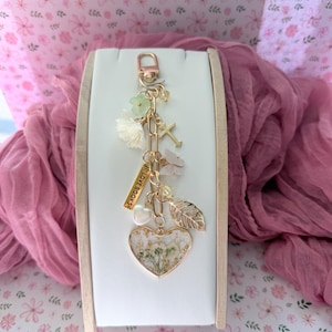 May include: A gold-tone keychain with a heart-shaped pendant filled with dried flowers, a cross, and a leaf charm. The keychain also includes small white and green floral accents and a charm that reads "LOVE BOOKS."