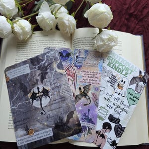May include: A collection of decorative bookmarks with fantasy-themed illustrations and text. The bookmarks feature images of dragons, castles, and quotes. White roses and an open book are in the background.