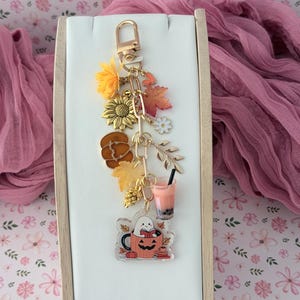 May include: A gold-toned metal keyring featuring a collection of autumn-themed charms including a sunflower, orange pumpkin, maple leaves, a pinecone, a small heart, a white flower, a boba tea cup, and a clear acrylic charm of a ghost in a pumpkin mug.