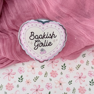 May include: A heart-shaped acrylic pin with a scalloped edge featuring the text Bookish Girlie in dark brown script, accented by two small pink cherries and four-pointed stars, all set against a cream background with a pink heart border.