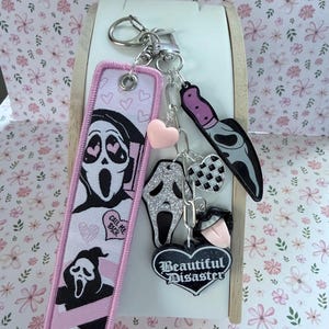 Pode incluir: Um chaveiro com vários berloques, incluindo um marcador rosa e preto com um gráfico de máscara Scream e o texto "Call Me Back". Outros berloques incluem uma faca, um coração e um coração "Beautiful Disaster".