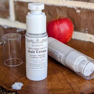 May include: A white bottle with a pump dispenser containing a lightweight leave-in hair cream. The bottle is labelled "Apothestix Lightweight Leave-in Hair Cream" and lists the product's benefits as "Smooth - Detangle - Condition - Protect". The bottle is sitting on a wooden surface with a red apple and a clear plastic bottle in the background.