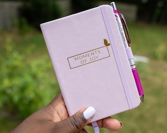 May include: A light purple notebook with the words "MOMENTS OF JOY" in gold lettering. A gold butterfly graphic is above the text. A silver and pink pen is tucked into the notebook's spine. The notebook is held in a hand.