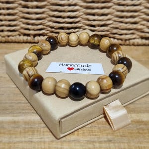 Puede incluir: Una pulsera de cuentas con cuentas redondas alternas de ojo de tigre pulido, madera de color tostado claro con veta visible y piedra negra sólida. Una pequeña etiqueta rectangular blanca tiene el texto Handmade with love en el centro.